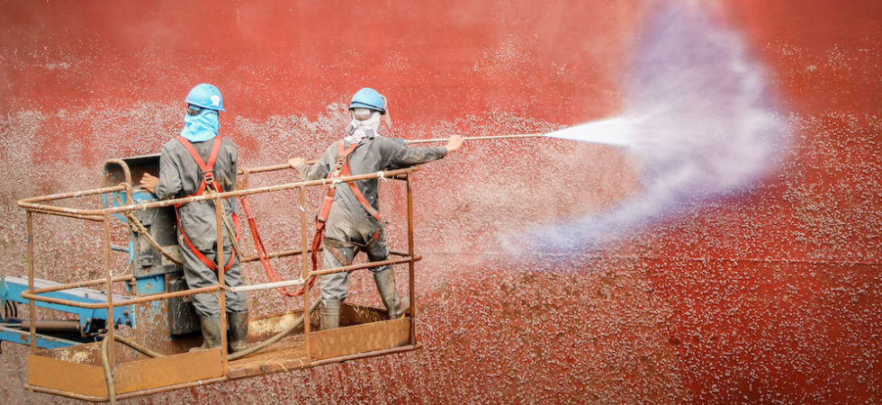 workers spraying the side of a ship with high pressure water to remove barnacles